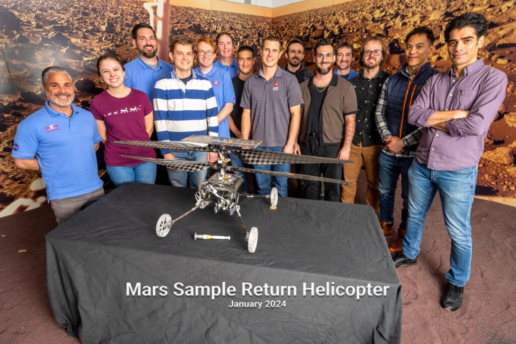 Color group photo of Hope Chambers on the left — wearing a maroon t-shirt with denim jeans — standing amongst 13 fellow male NASA JPL colleagues with the Mars Sample Return Helicopter displayed on a table with a black tablecloth. Text reads: “Mars Sample Return Helicopter | January 2024"